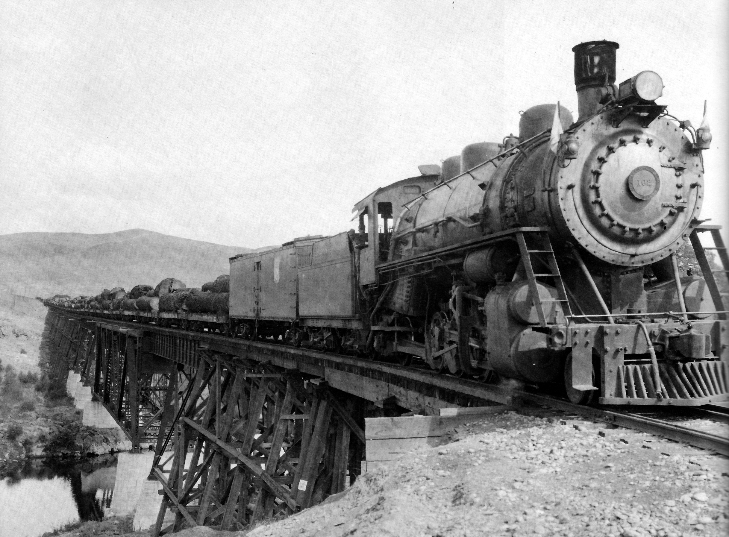 A black-and-white photograph of a steam locomotive hauling a freight train across a wooden trestle bridge.
